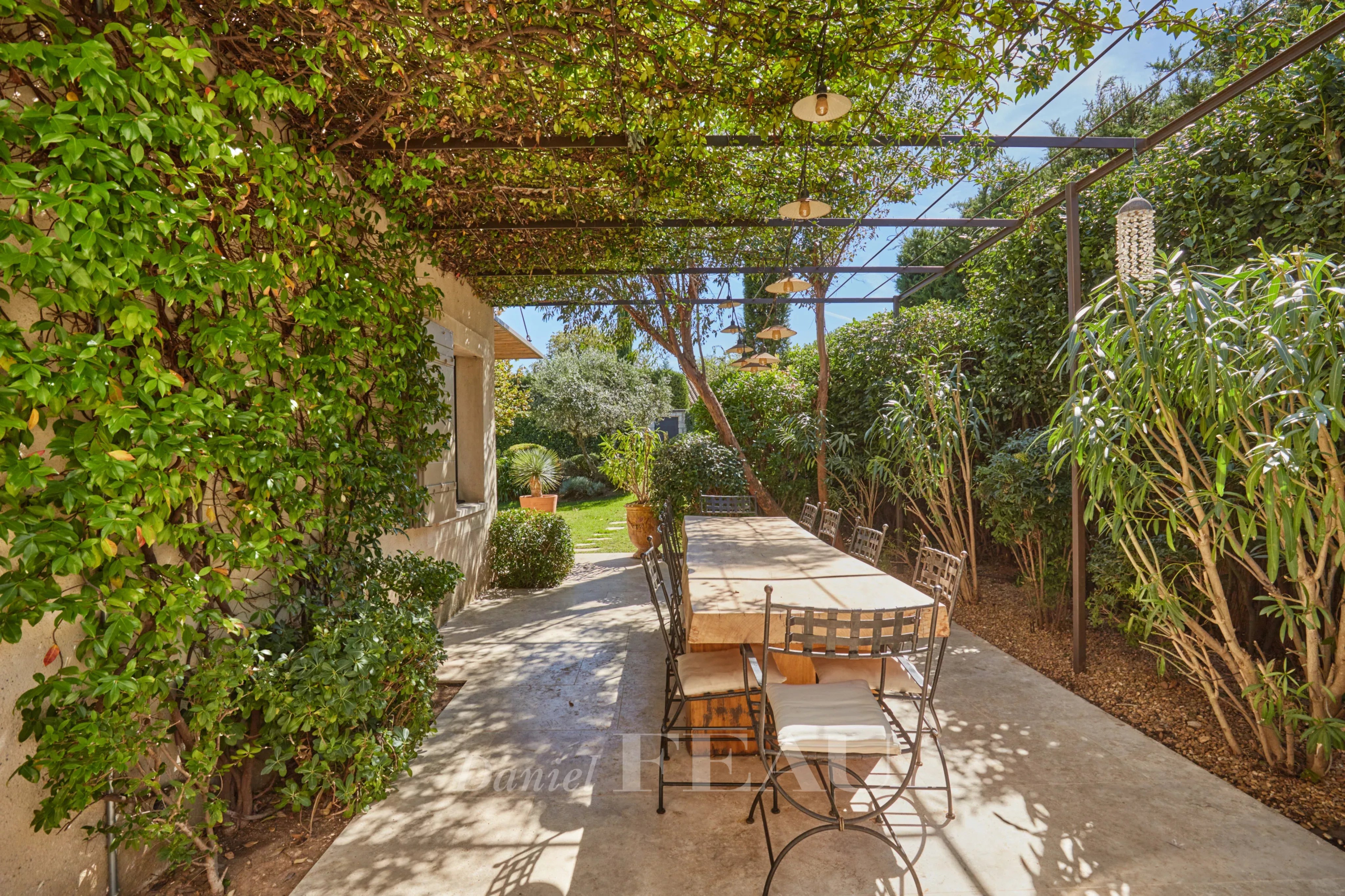 Terrasse d’une maison à Eygalières Terrasse d'une maison à Eygalières avec une table en bois et des chaises, entourée de verdure.