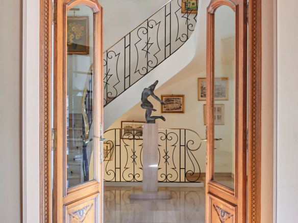 Open double wooden doors frame a gallery view: a marble floor, a pedestal sculpture, and a curving wrought-iron staircase in the background.