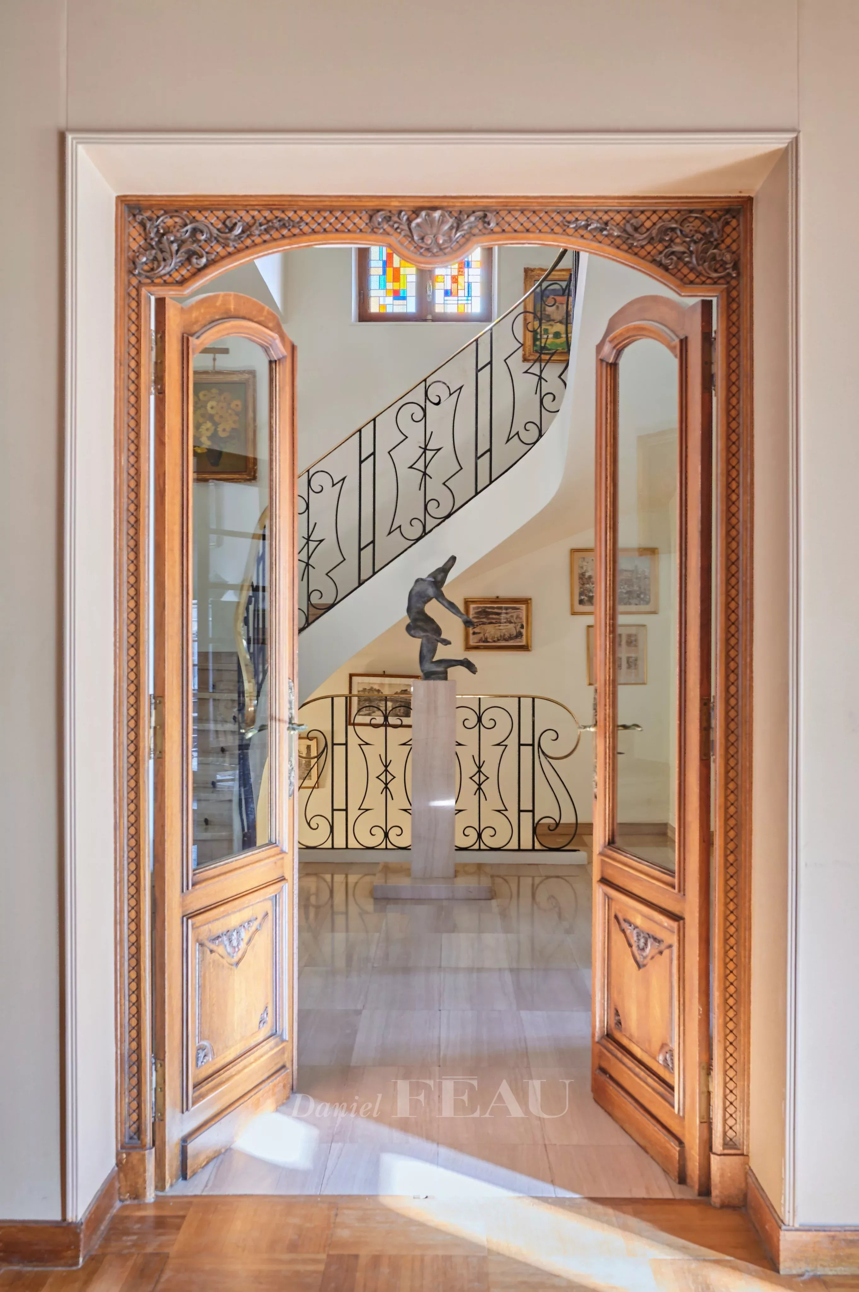 Open double wooden doors frame a gallery view: a marble floor, a pedestal sculpture, and a curving wrought-iron staircase in the background.