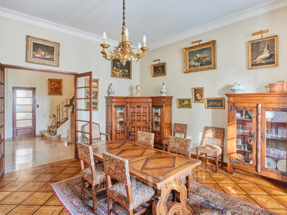 Antique wooden dining room with a large parquet table surrounded by upholstered chairs, a chandelier overhead, and glass-front cabinets along the walls displaying china and decor.