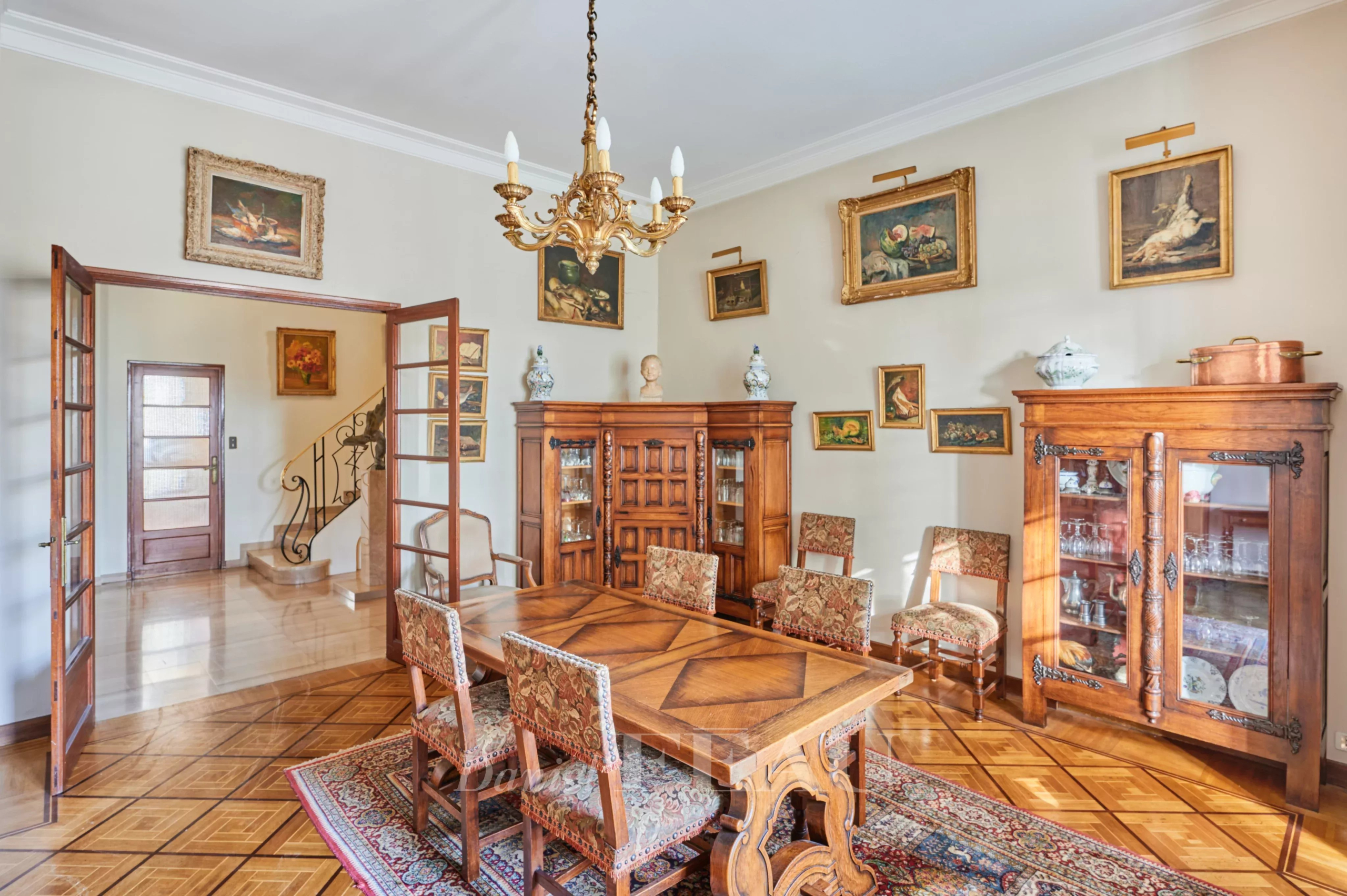 Antique wooden dining room with a large parquet table surrounded by upholstered chairs, a chandelier overhead, and glass-front cabinets along the walls displaying china and decor.