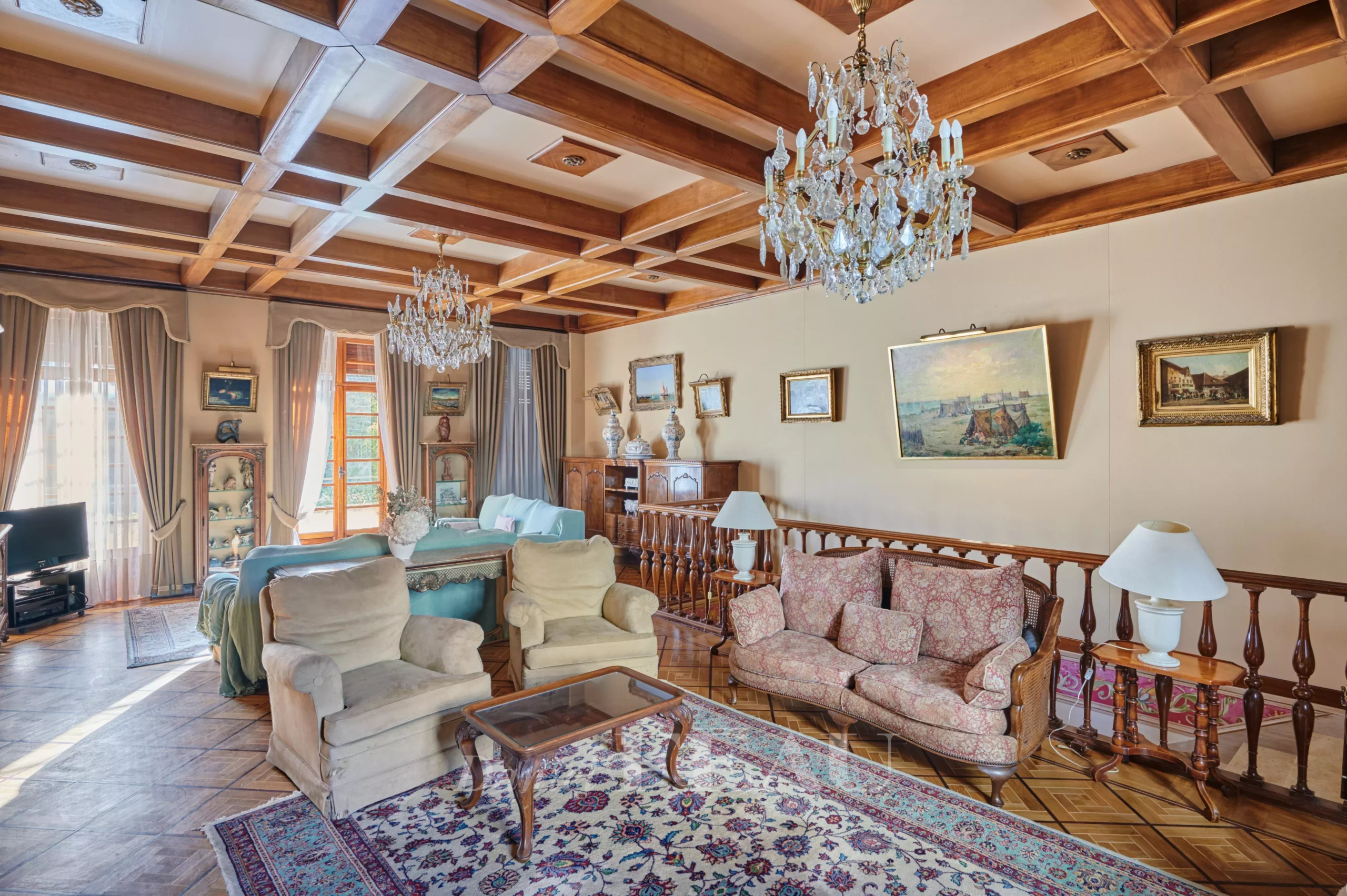 Luxurious living room with a coffered wooden ceiling, crystal chandeliers, and antique seating including a floral pink sofa and beige armchairs.