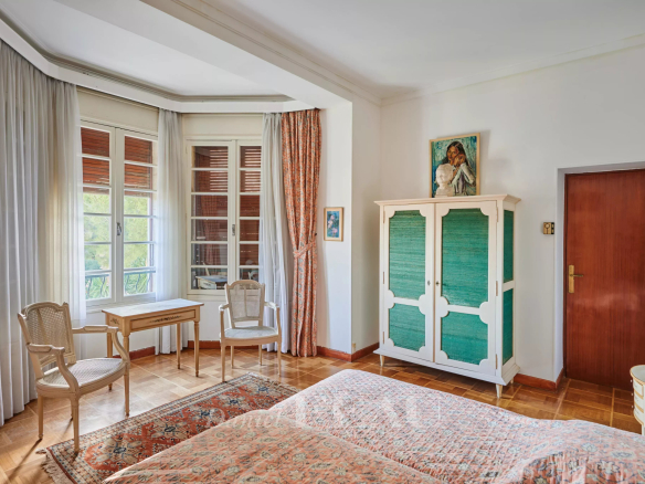 Bright vintage bedroom with a bay window, sheer white curtains, two wicker chairs and a small wooden desk by the window.