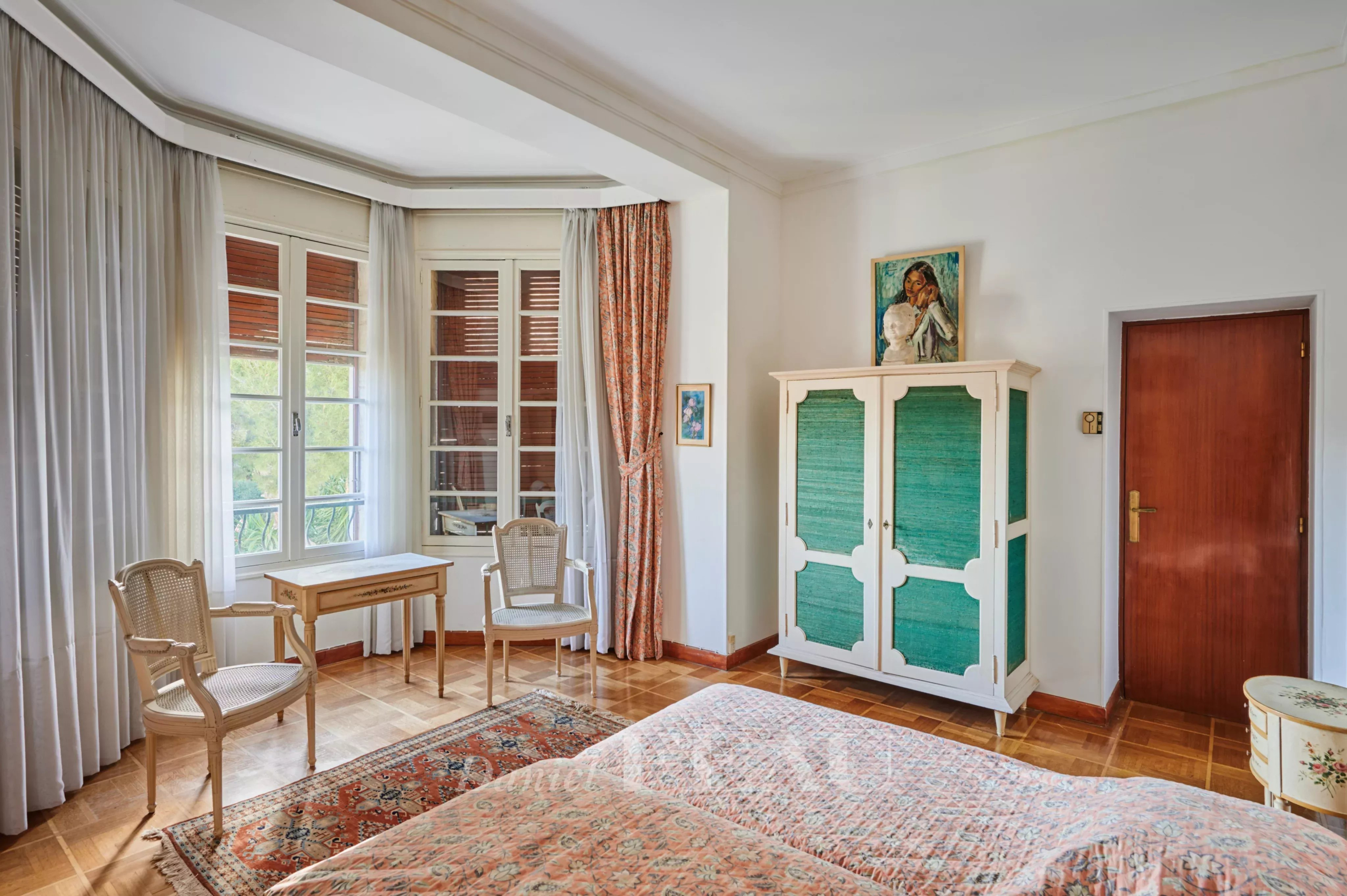 Bright vintage bedroom with a bay window, sheer white curtains, two wicker chairs and a small wooden desk by the window.