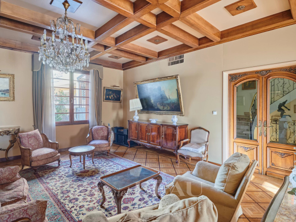 Elegant traditional living room with a crystal chandelier, wooden beam ceiling, and antique furniture including armchairs and a sideboard.