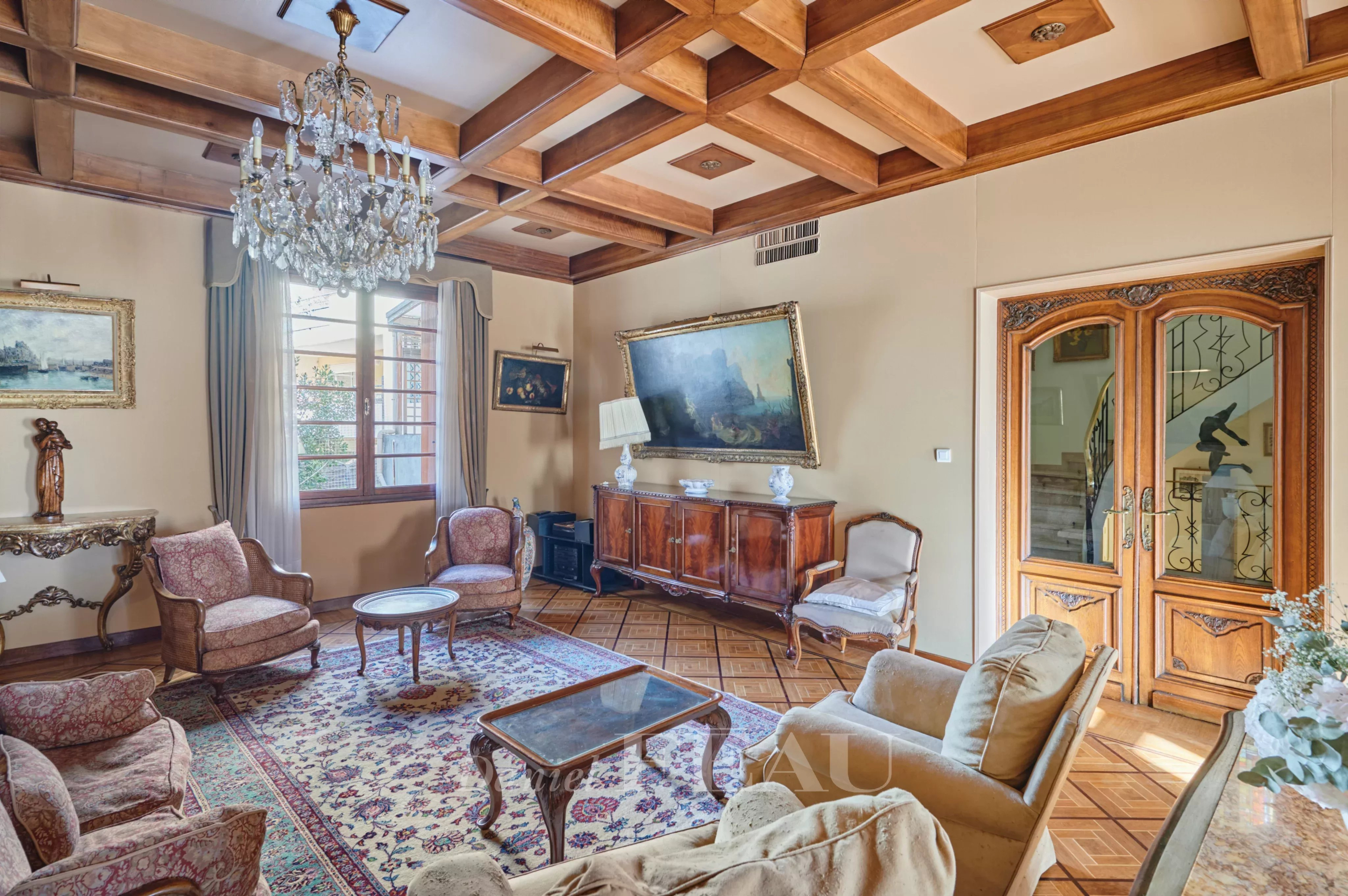 Elegant traditional living room with a crystal chandelier, wooden beam ceiling, and antique furniture including armchairs and a sideboard.