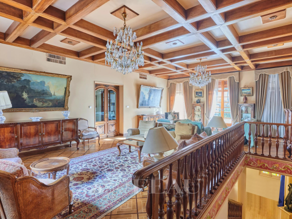 Luxurious living room with a coffered wooden ceiling, crystal chandeliers, and antique wood furniture surrounding a patterned rug.