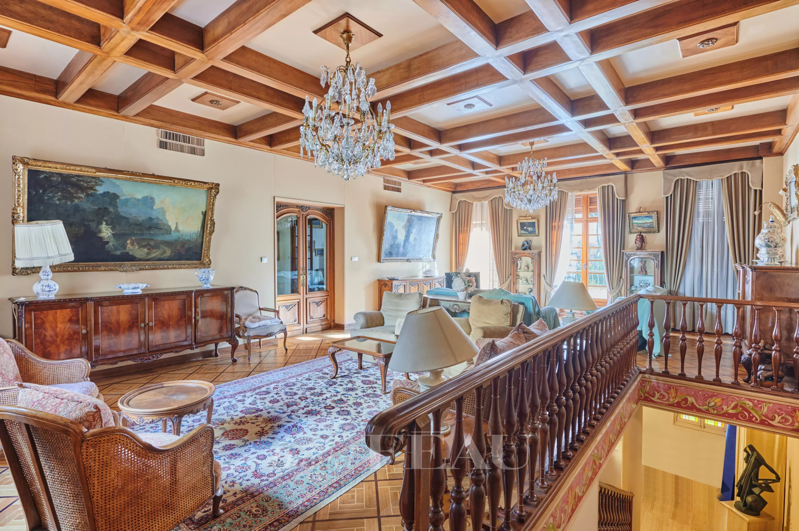 Luxurious living room with a coffered wooden ceiling, crystal chandeliers, and antique wood furniture surrounding a patterned rug.