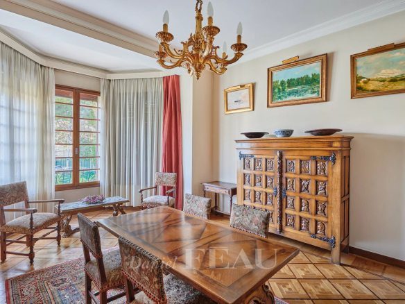 Elegant dining room with antique wooden table and carved chairs, a brass chandelier, and a large carved sideboard; sunlit window with sheer curtains and framed landscape paintings above the sideboard.