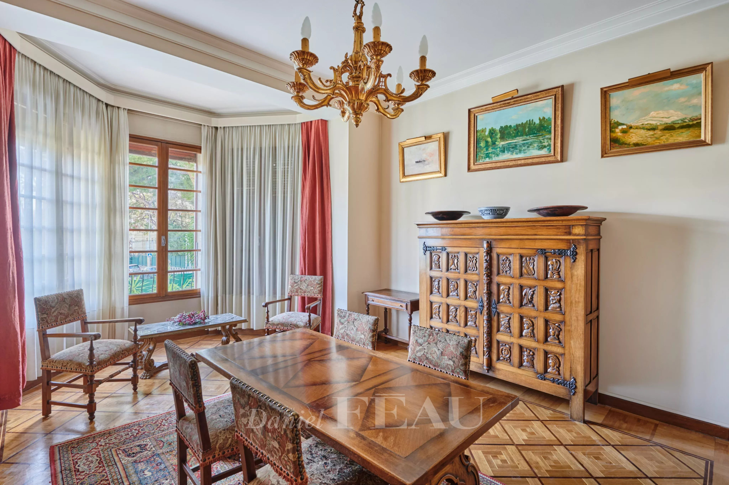 Elegant dining room with antique wooden table and carved chairs, a brass chandelier, and a large carved sideboard; sunlit window with sheer curtains and framed landscape paintings above the sideboard.
