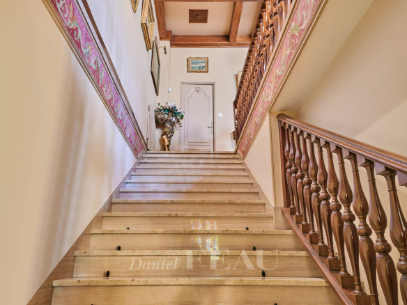 Grand interior staircase with carved wooden railing, pink decorative border, and framed art along the walls leading to a door. The landing features a vase with flowers near the door at top of stairs.