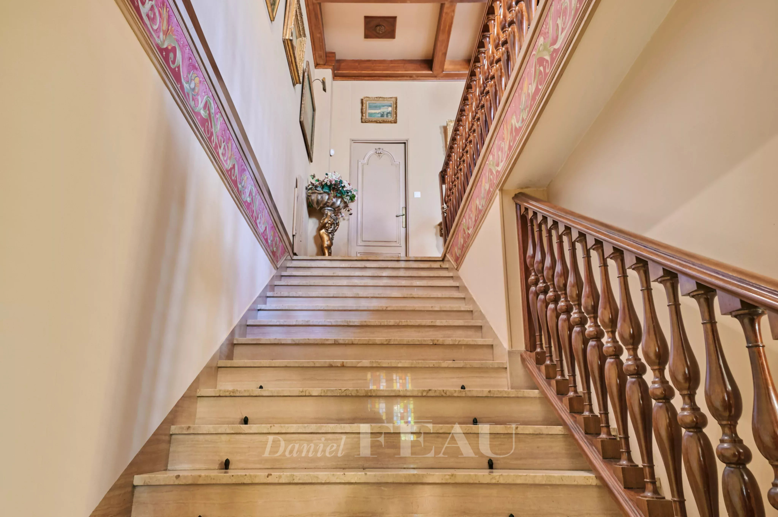 Grand interior staircase with carved wooden railing, pink decorative border, and framed art along the walls leading to a door. The landing features a vase with flowers near the door at top of stairs.