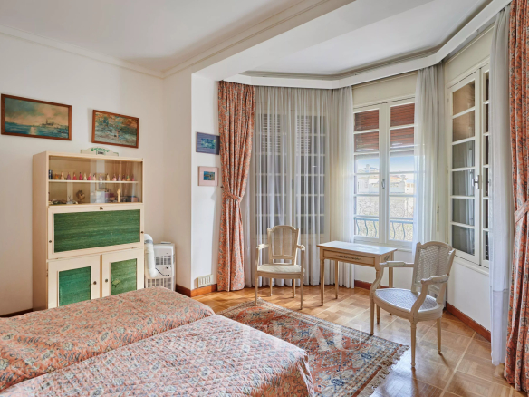 Sunlit vintage bedroom with two twin beds, patterned quilts, and a writing desk near large bay windows.