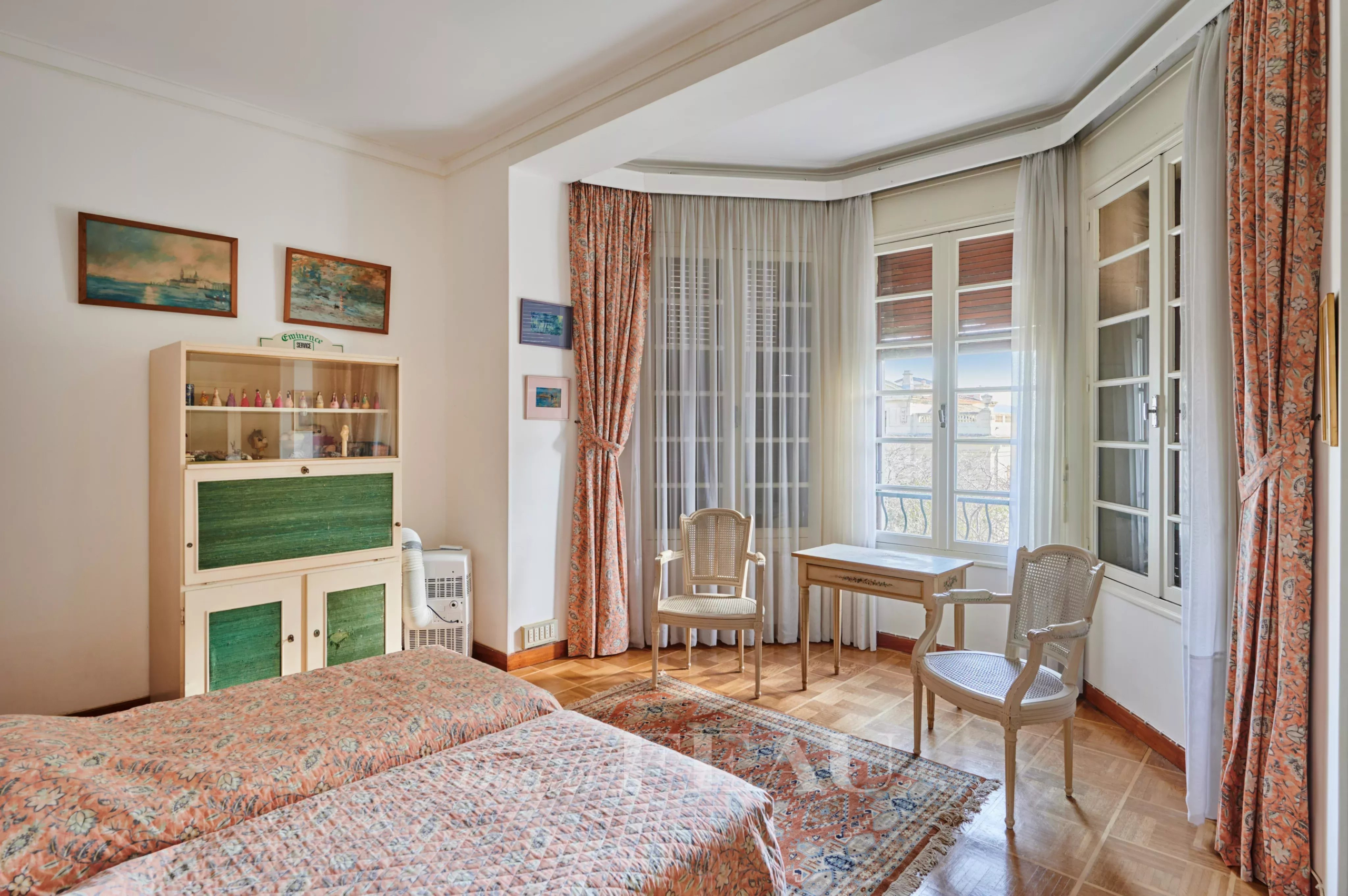 Sunlit vintage bedroom with two twin beds, patterned quilts, and a writing desk near large bay windows.