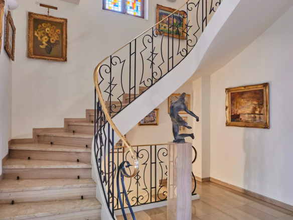 Curved staircase with ornate black iron railing, a statue on a pedestal, and framed paintings in a bright foyer.
