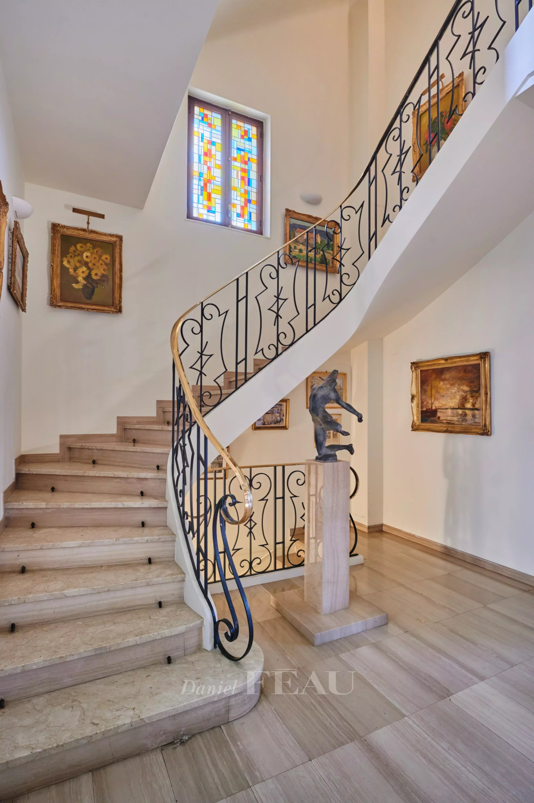 Curved staircase with ornate black iron railing, a statue on a pedestal, and framed paintings in a bright foyer.