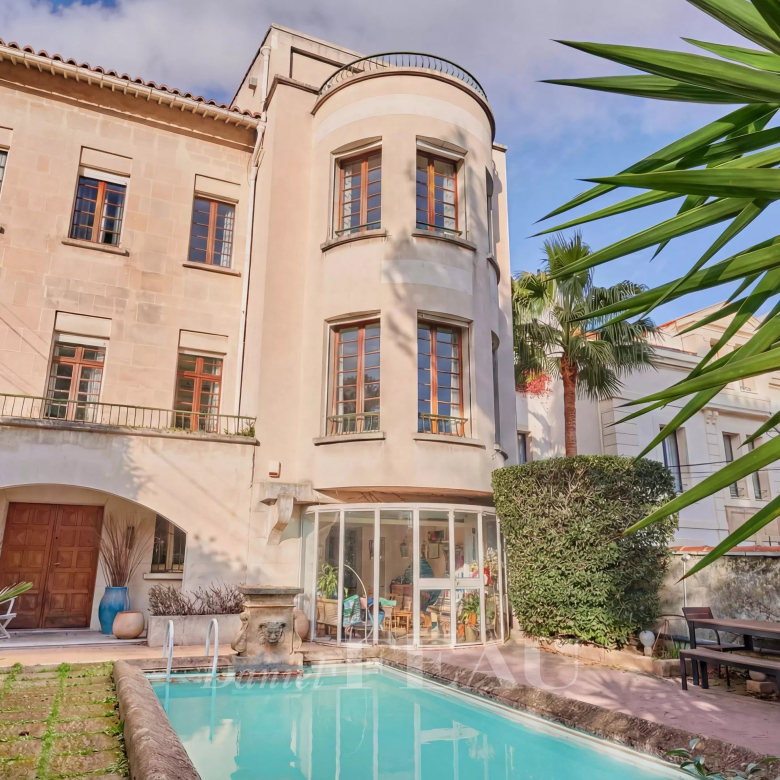 Mediterranean-style villa with a curved bay window, glass conservatory, and a rectangular pool in a sunny courtyard surrounded by plants.