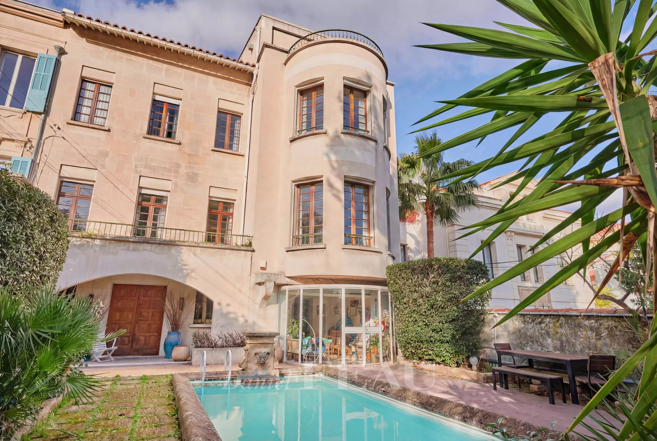 Mediterranean-style villa with a curved bay window, glass conservatory, and a rectangular pool in a sunny courtyard surrounded by plants.