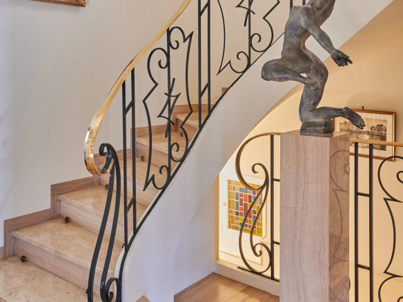 Elegant spiral staircase with ornate black iron railing and a sculpted figure on a marble pedestal at the base, surrounded by framed paintings on light walls.
