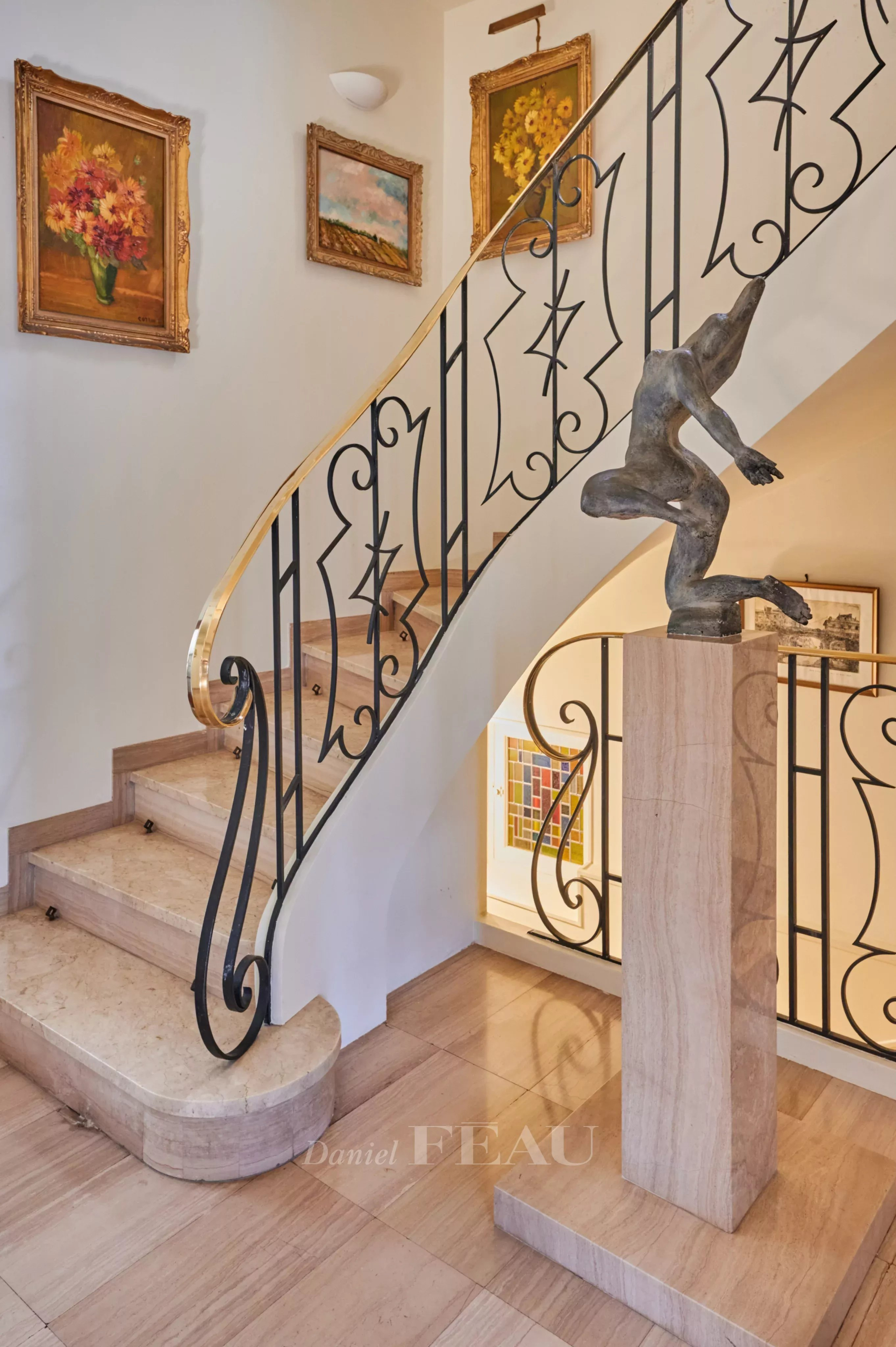 Elegant spiral staircase with ornate black iron railing and a sculpted figure on a marble pedestal at the base, surrounded by framed paintings on light walls.
