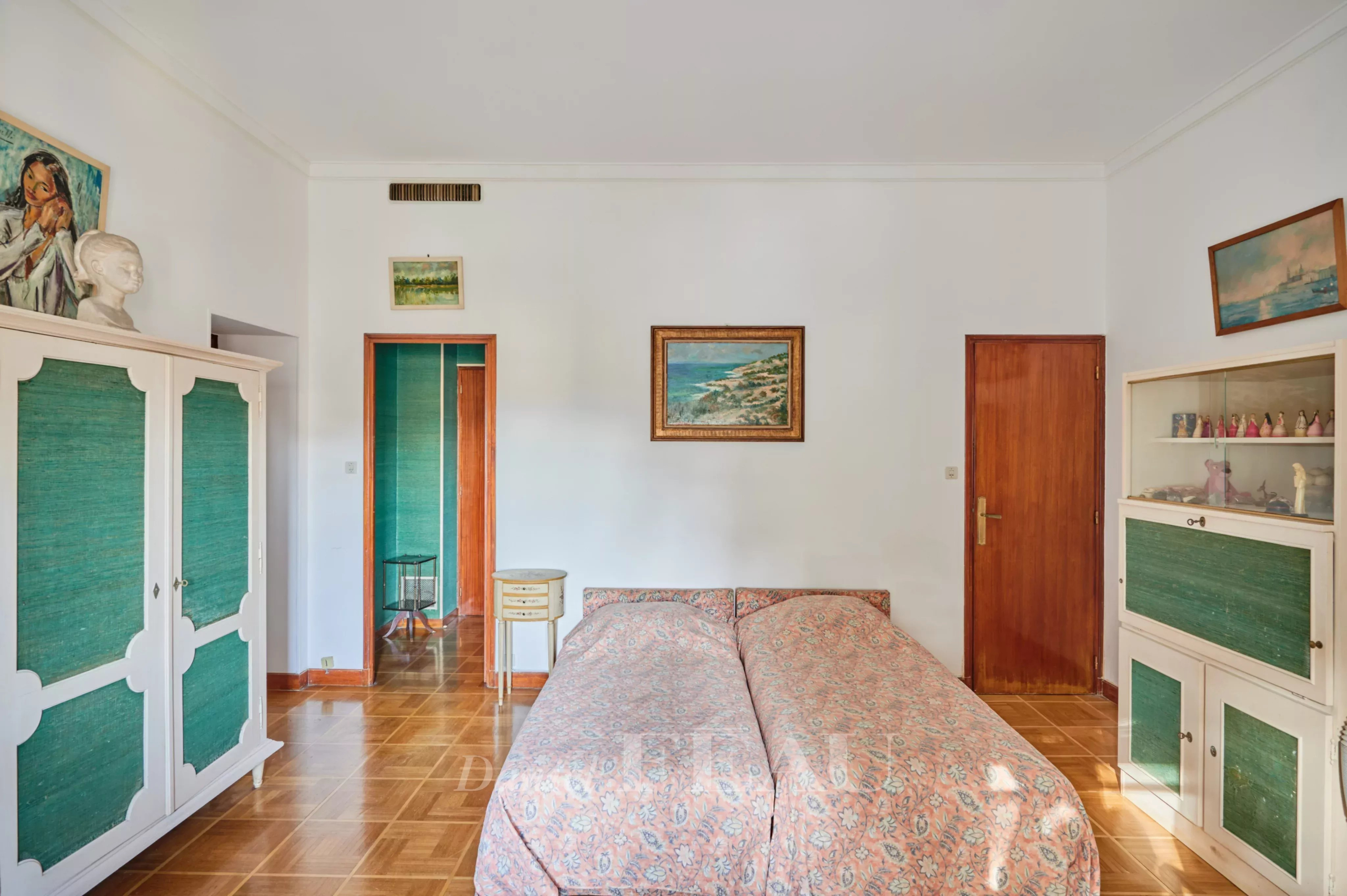 Twin beds with pink floral covers in a vintage bedroom, flanked by white and green-paneled wardrobes and wooden doors.