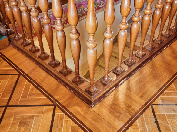 Wooden staircase railing with carved balusters and a polished handrail, seen over a parquet floor with geometric inlay patterns