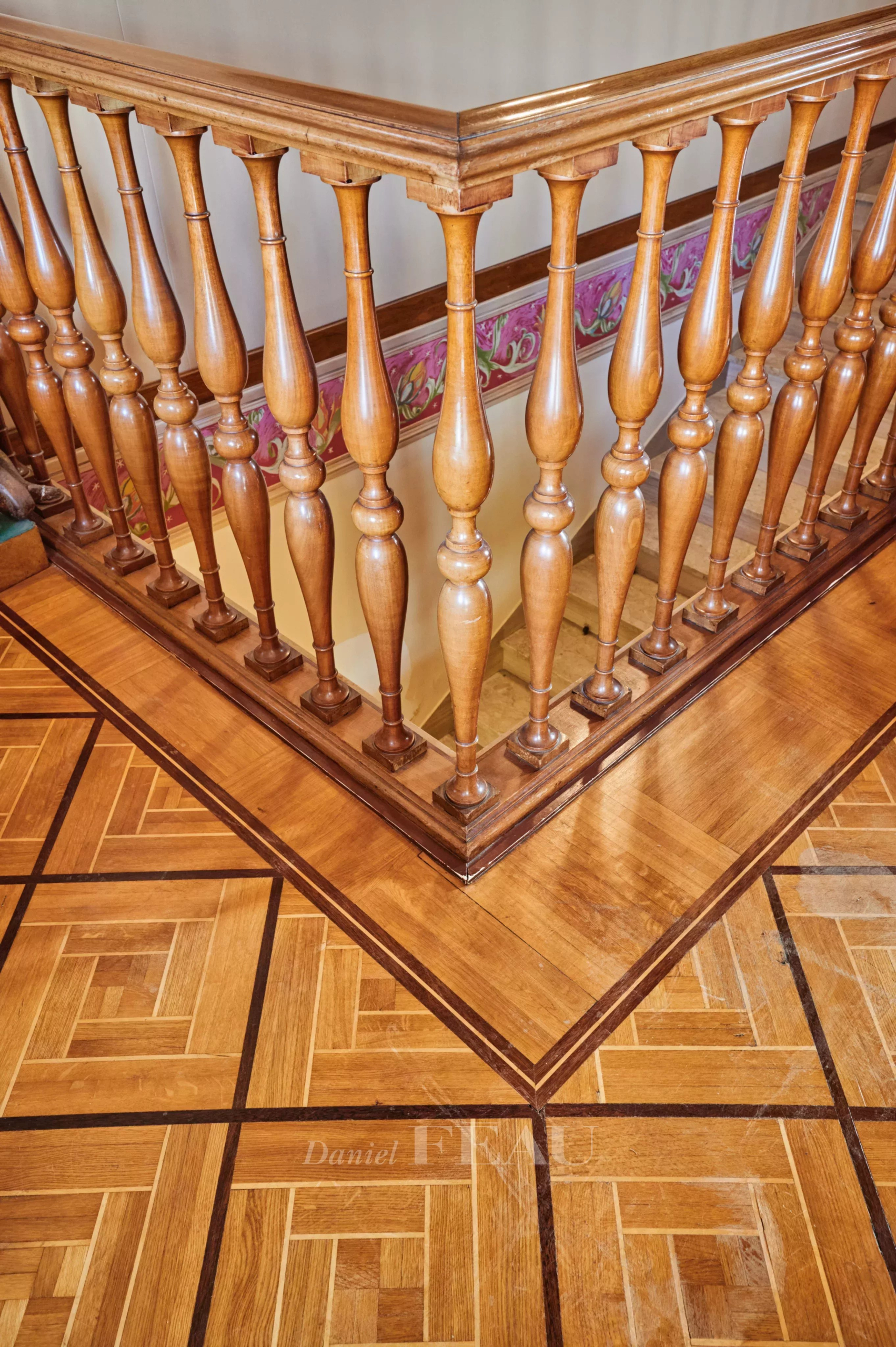 Wooden staircase railing with carved balusters and a polished handrail, seen over a parquet floor with geometric inlay patterns