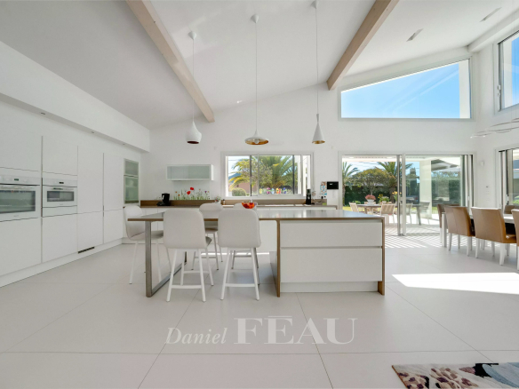 Bright, modern white kitchen with an island and white stools, open to a sunny patio through large doors.