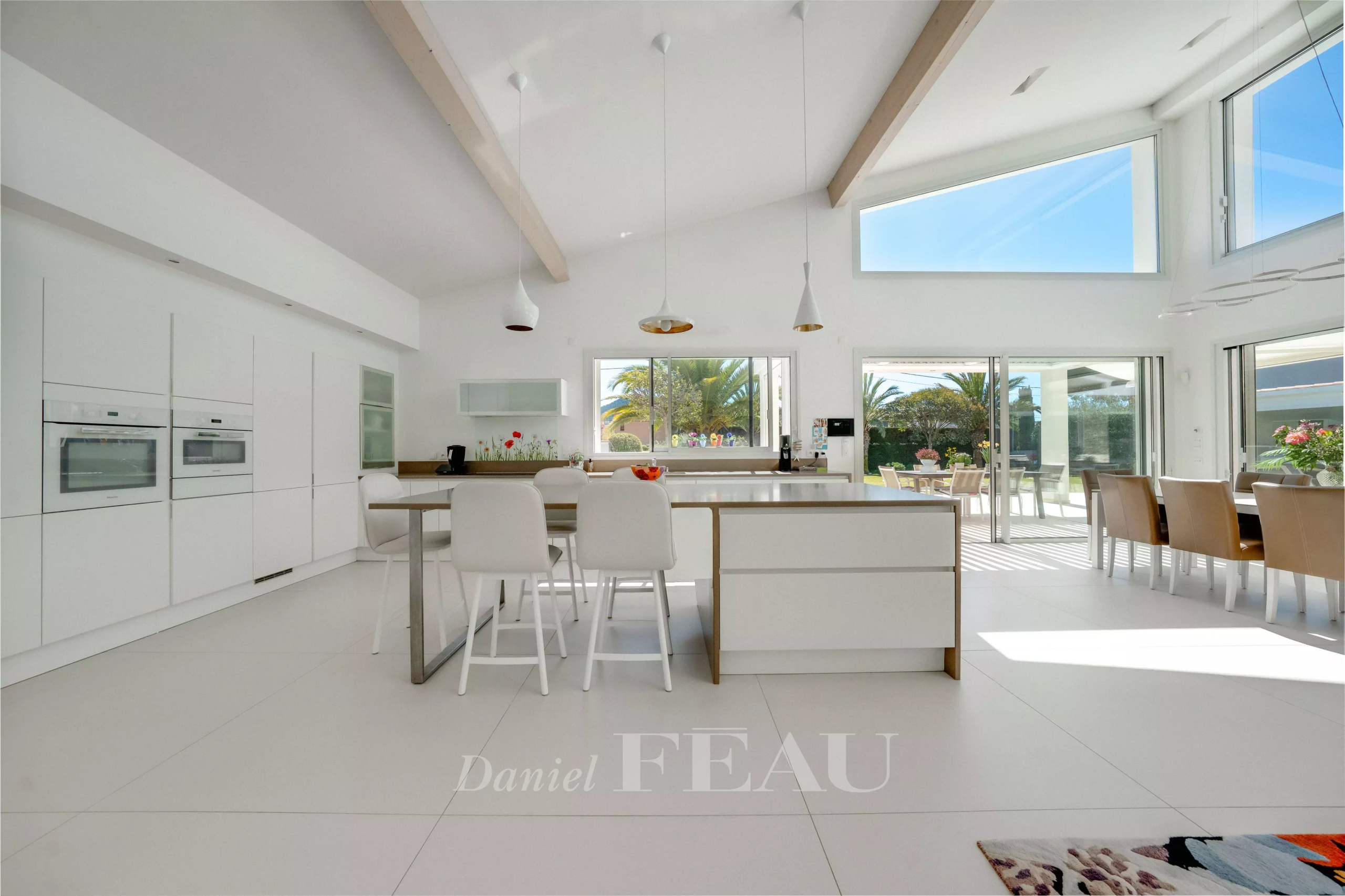 Bright, modern white kitchen with an island and white stools, open to a sunny patio through large doors.