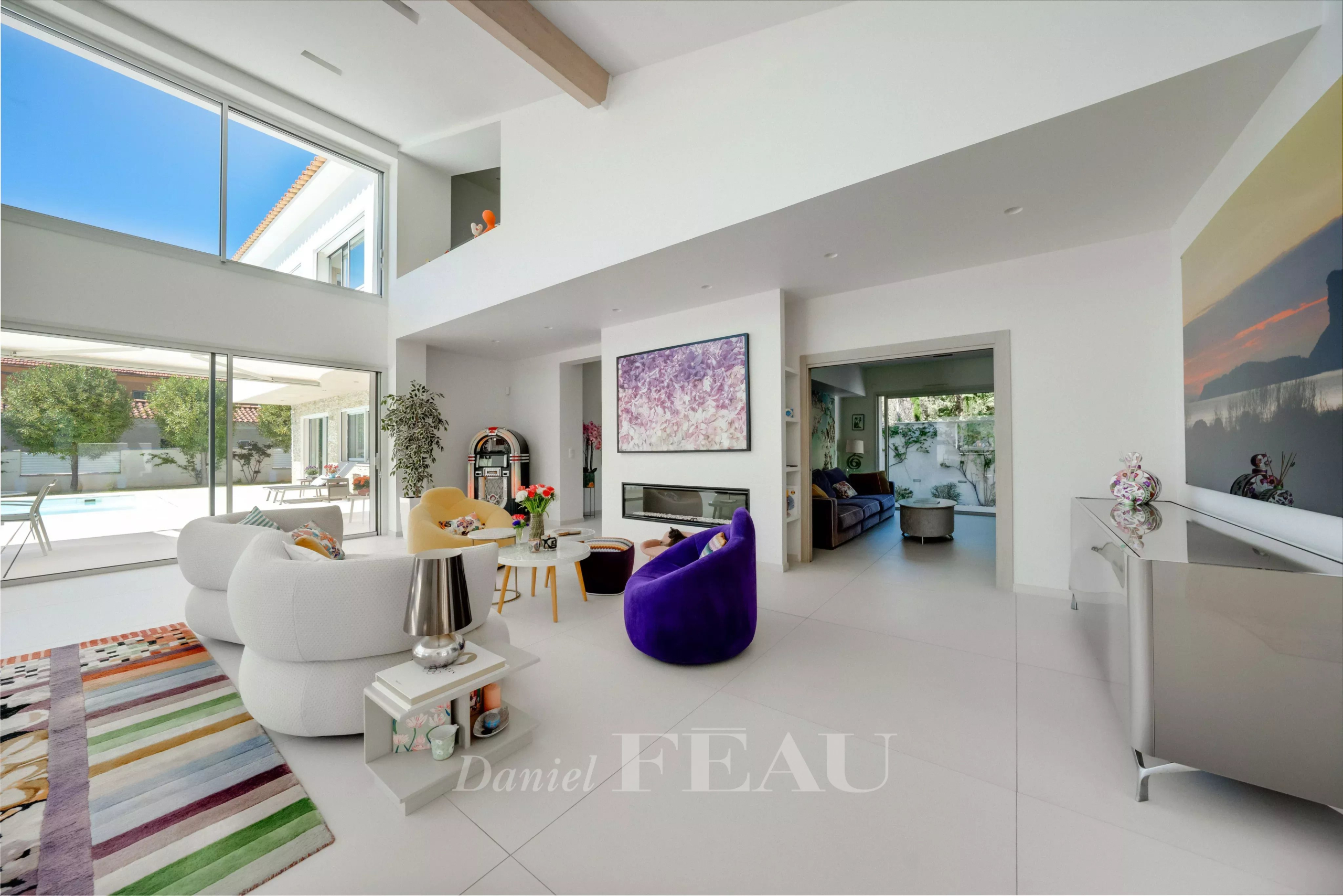 Bright open living room with white furniture, a purple chair, colorful rug, fireplace, and large glass walls showing an outdoor pool.