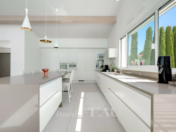 Bright, modern white kitchen with a long island, built-in ovens, and a large window showing green trees outside