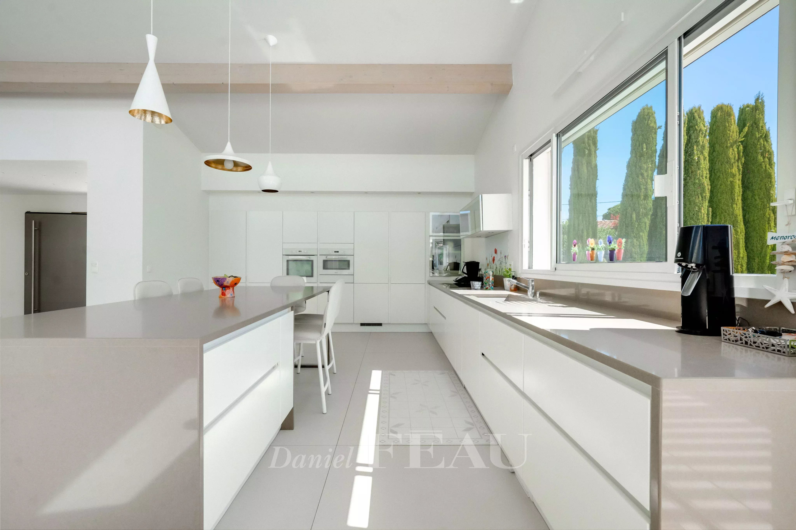 Bright, modern white kitchen with a long island, built-in ovens, and a large window showing green trees outside