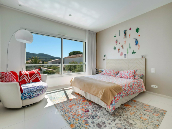 Bright bedroom with a large window overlooking hills, a white curved lounge chair with red cushions, and a colorful tropical-themed bed setup.
