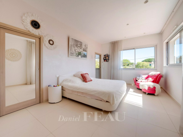 Bright, sunlit bedroom with a white bed, fluffy duvet, and pink throw pillows by a large windowed wall.