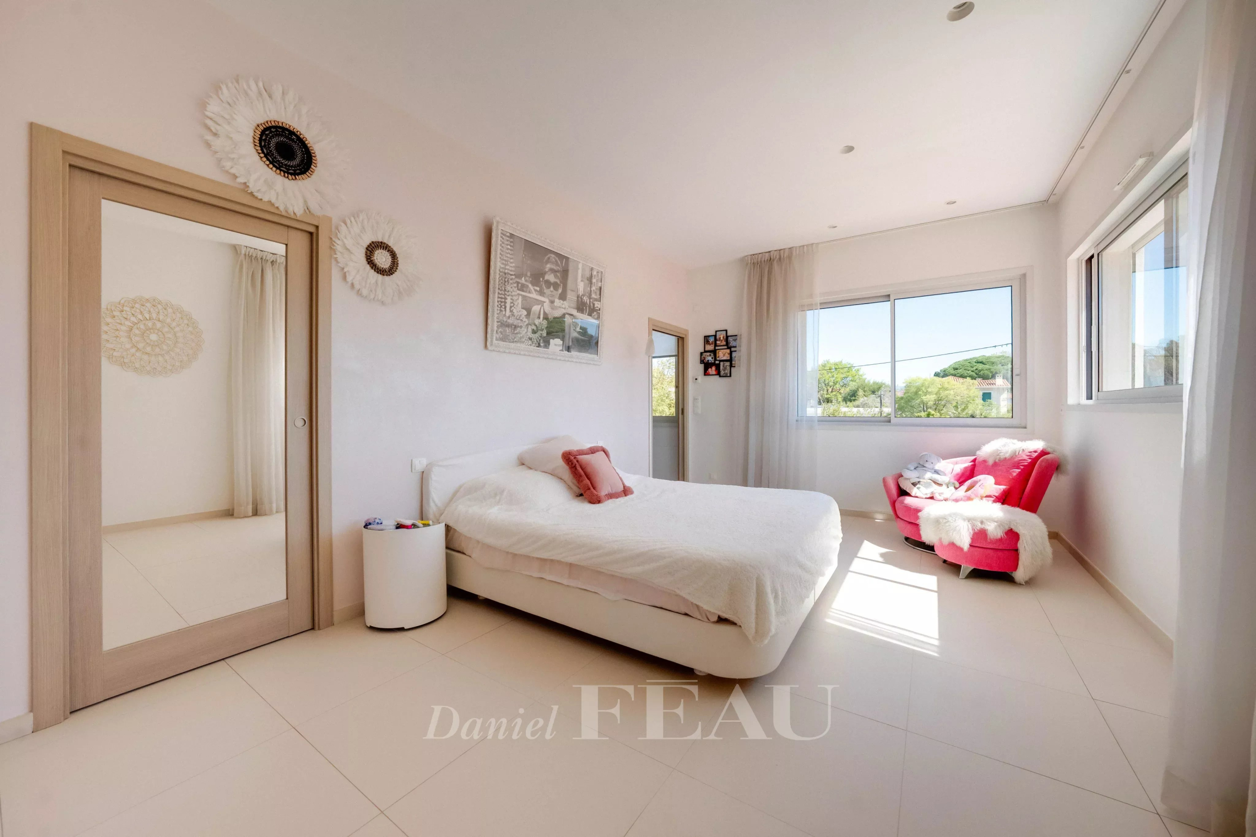 Bright, sunlit bedroom with a white bed, fluffy duvet, and pink throw pillows by a large windowed wall.