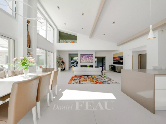 Bright open-plan living and dining area with white furniture, beige chairs, and a colorful rug bathed in natural light from large windows on the left.
