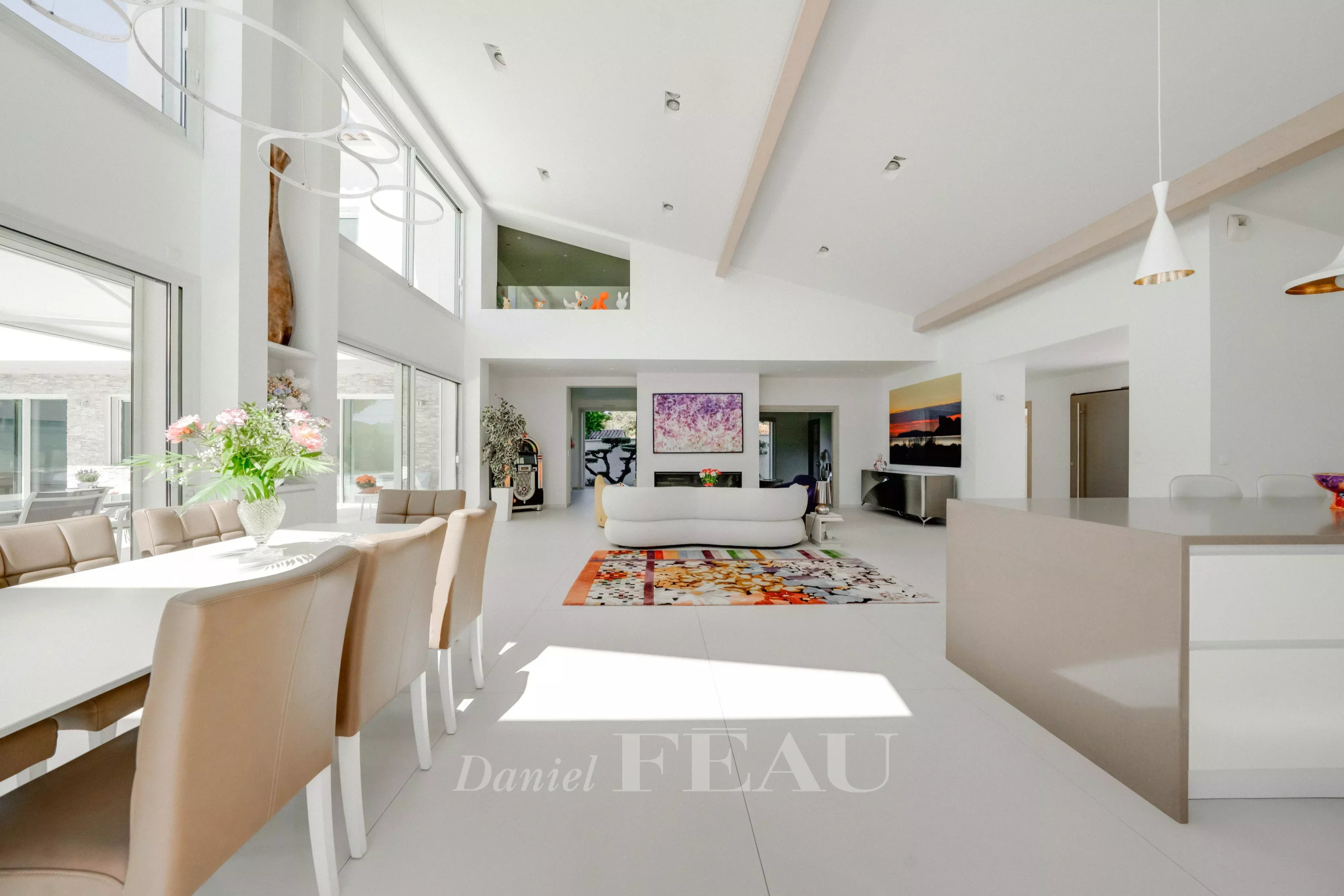 Bright open-plan living and dining area with white furniture, beige chairs, and a colorful rug bathed in natural light from large windows on the left.