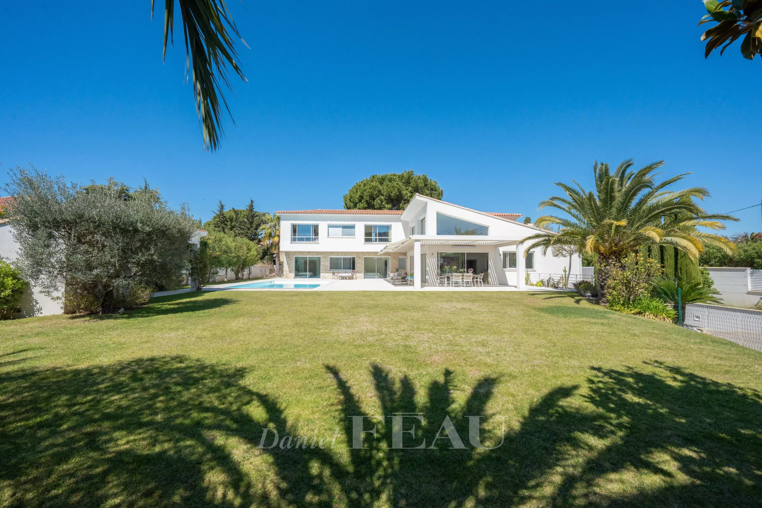 White contemporary two-story house with a rectangular pool in a spacious lawn and palm trees under a clear blue sky.
