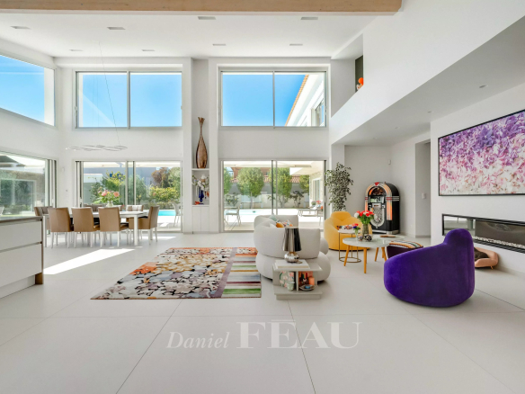 Bright open living room with white floors and large windows, colorful seating, and a dining area by glass walls overlooking a backyard pool.