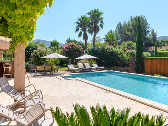 View of a property with swimming pool, sun loungers and palm trees, surrounded by greenery.