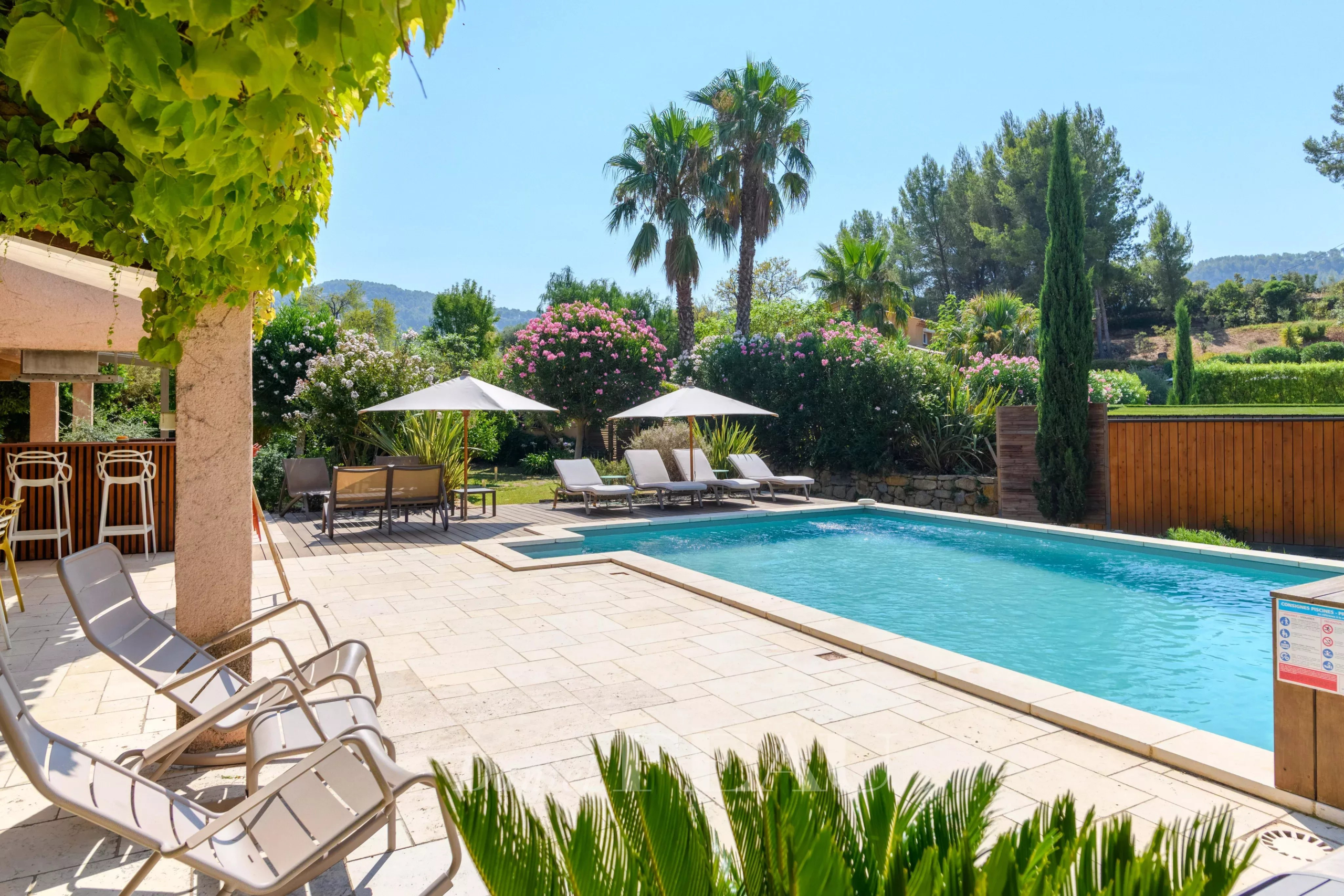 View of a property with swimming pool, sun loungers and palm trees, surrounded by greenery.