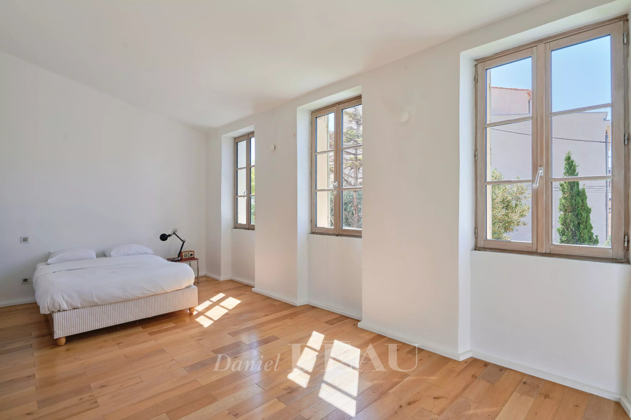 Chambre lumineuse avec fenêtres sur trois côtés, parquet en bois et lit simple.