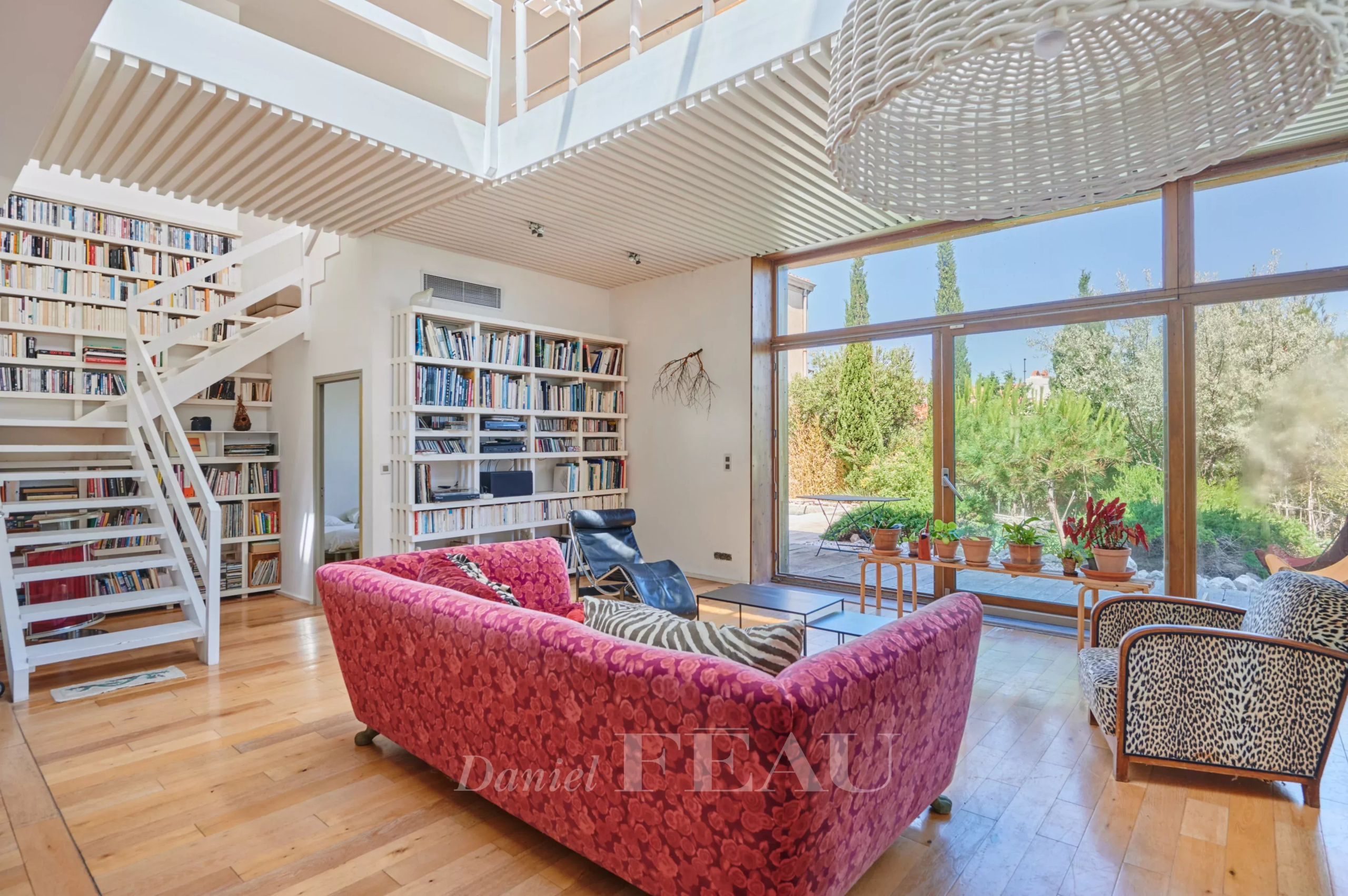 Intérieur d'une maison moderne avec un salon lumineux, des étagères et un jardin visible à travers les grandes fenêtres.