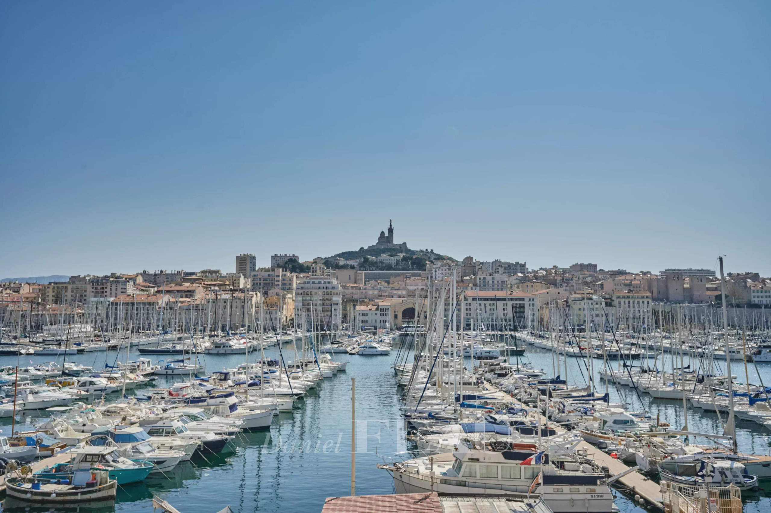 Vue panoramique du Vieux-Port de Marseille avec des bateaux à quai et la basilique Notre-Dame de la Garde en arrière-plan.