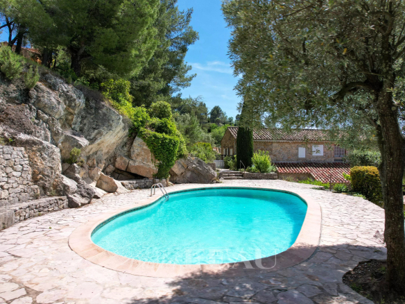 Piscine en forme libre entourée de rochers et de verdure, avec une bastide en pierre en arrière-plan.