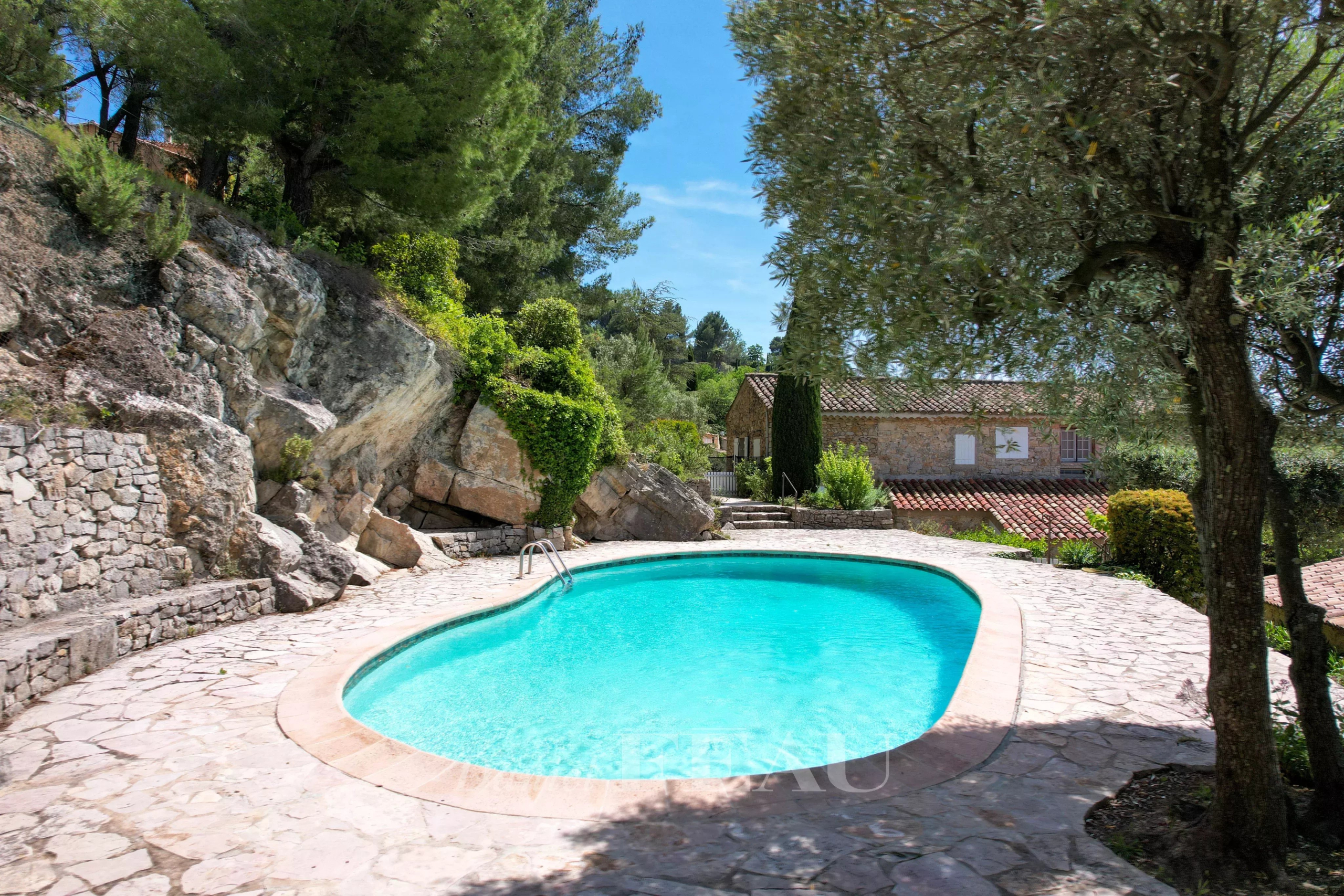 Piscine de la bastide au Beausset Piscine en forme libre entourée de rochers et de verdure, avec une bastide en pierre en arrière-plan.