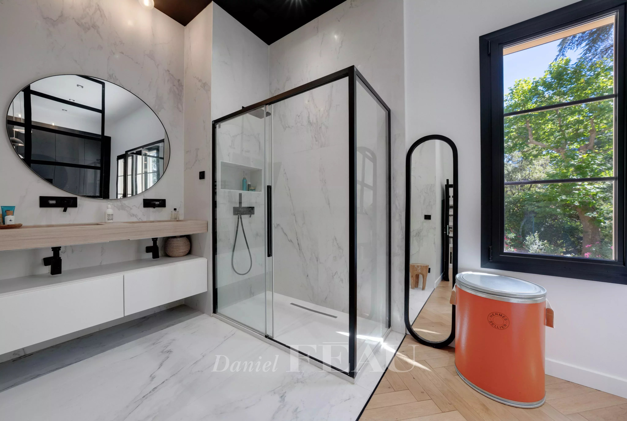 Salle de bain moderne avec douche à l'italienne et miroir rond, lumière naturelle à travers la fenêtre.