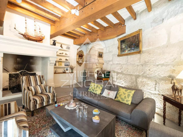 Cozy living room with a stone wall, wooden beams, and a white mantel featuring a ship model and shelves of small decor items behind striped armchairs and a gray sofa with patterned cushions.