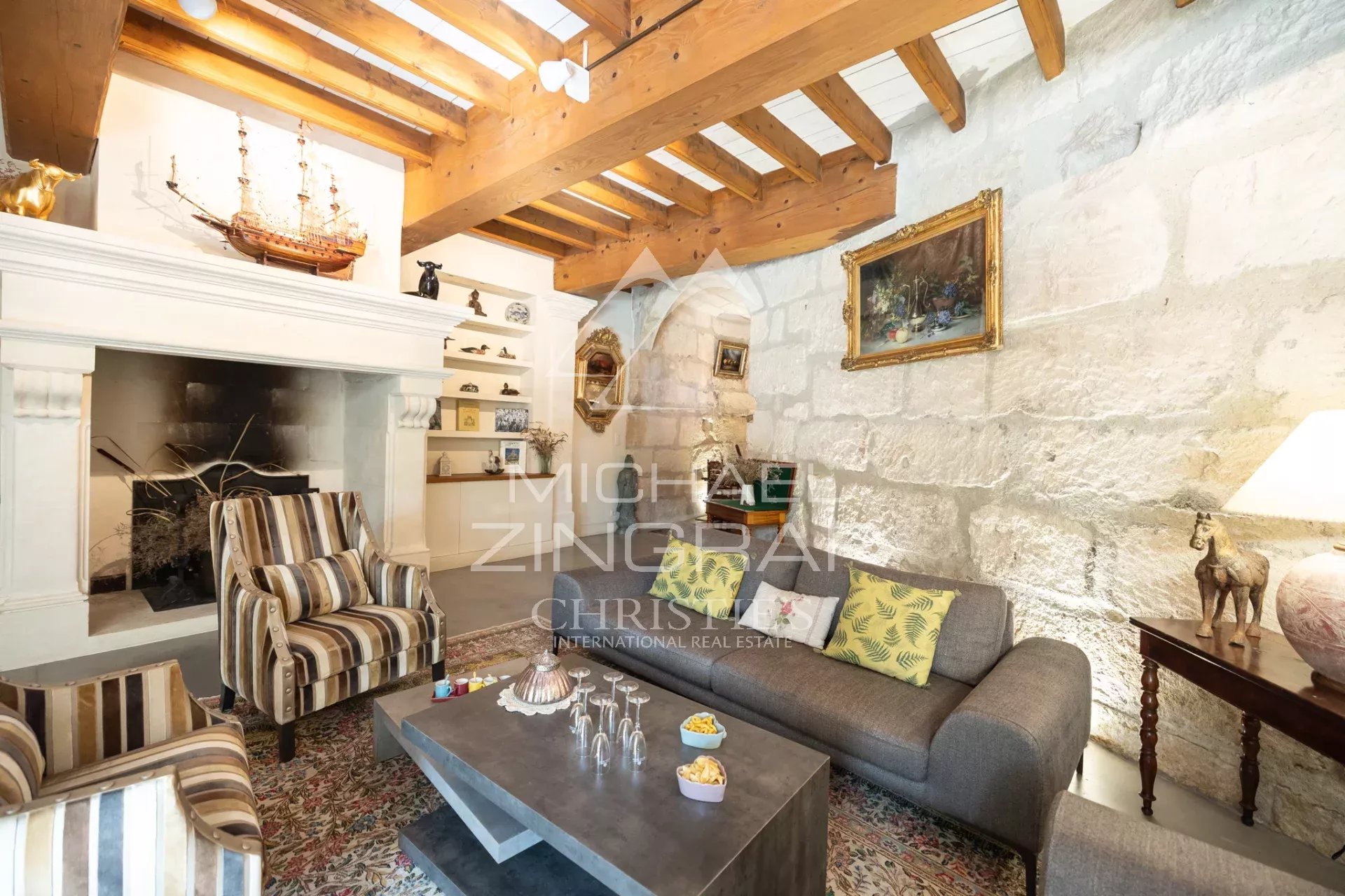 Cozy living room with a stone wall, wooden beams, and a white mantel featuring a ship model and shelves of small decor items behind striped armchairs and a gray sofa with patterned cushions.