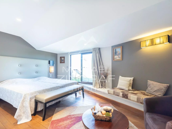 Spacious hotel bedroom with a king bed and padded headboard, a bench at the foot, window seating, and a round table with a basket of snacks nearby.
